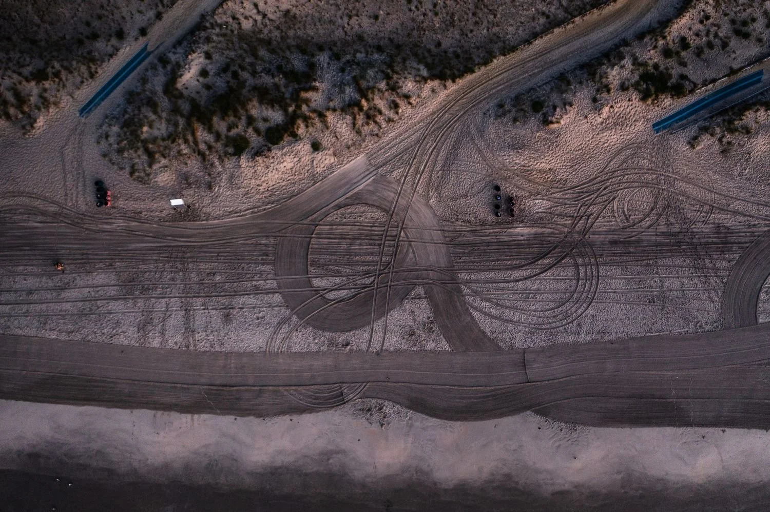 An aerial view of a dirt parking lot with multiple tire tracks, surrounded by some vegetation and a few parked cars.