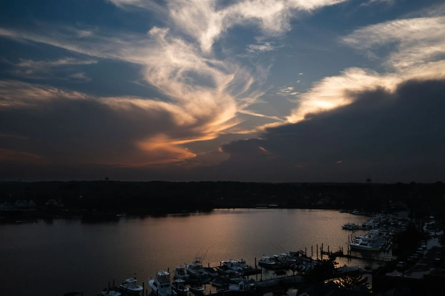 Sunset over a marina with boats docked along the water, dark clouds in the sky, and a reflective body of water in the foreground.