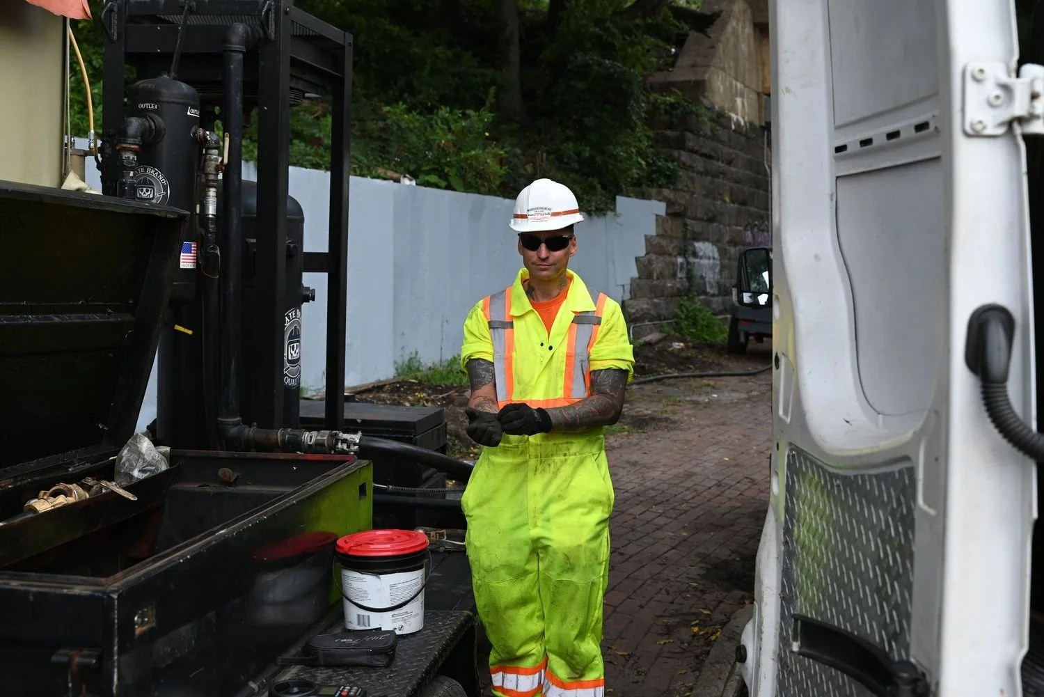 A worker in a yellow safety vest, neon green coveralls, a white hard hat, and sunglasses standing next to a utility truck with equipment and tools, with a brick wall and trees in the background.