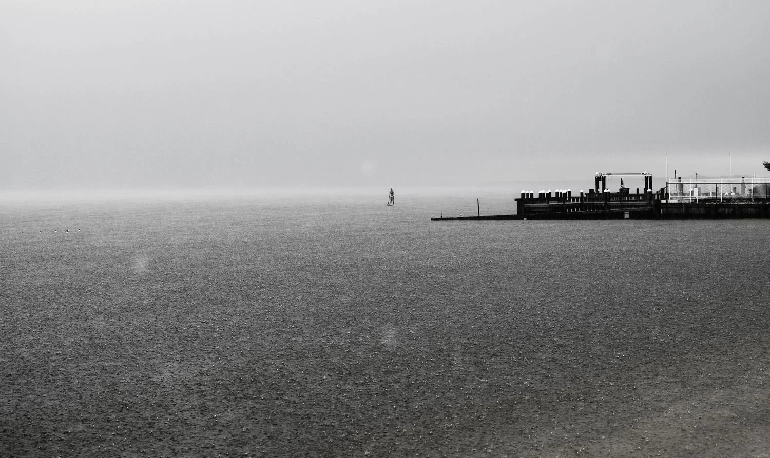 Black and white photograph of a calm body of water with a small pier on the right and a lone person standing on a platform in the distance.
