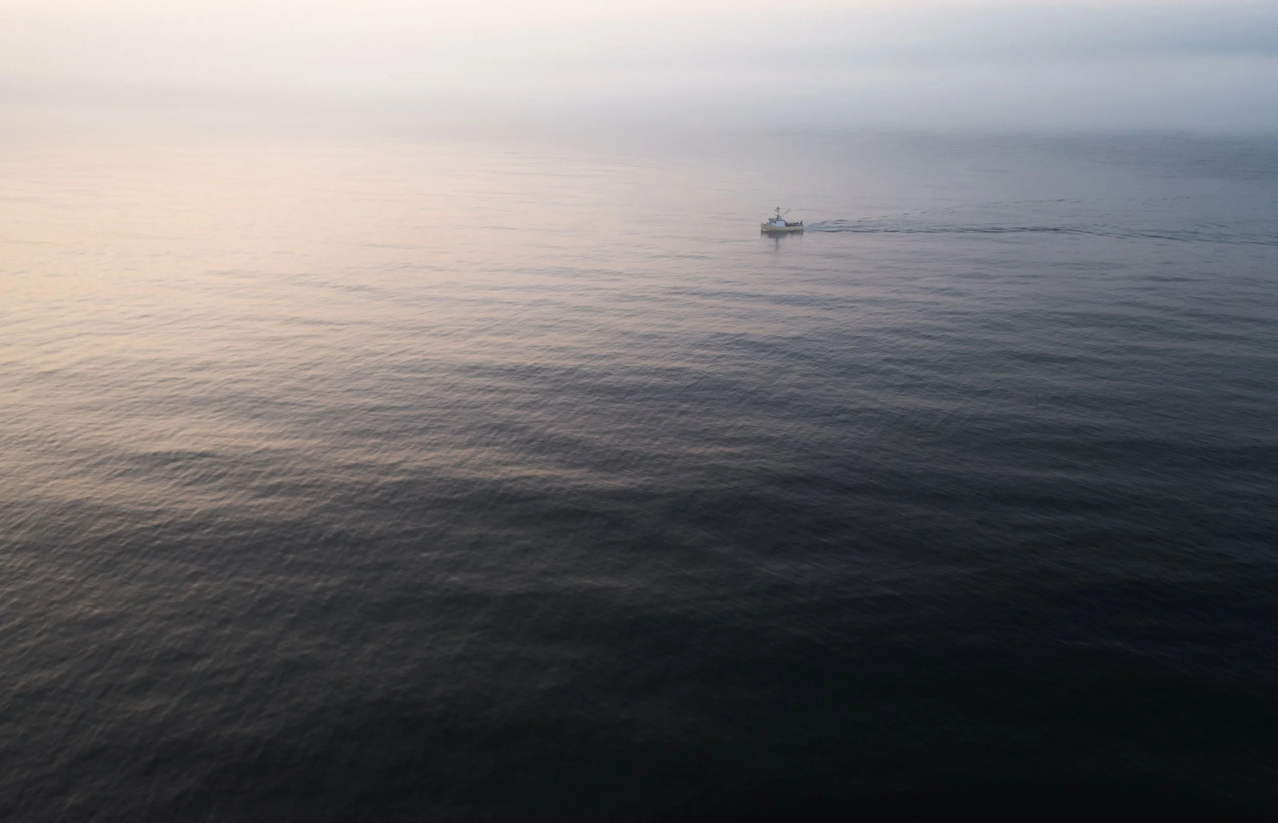 A small boat sailing on calm ocean waters near sunset or sunrise with a light gradient sky.