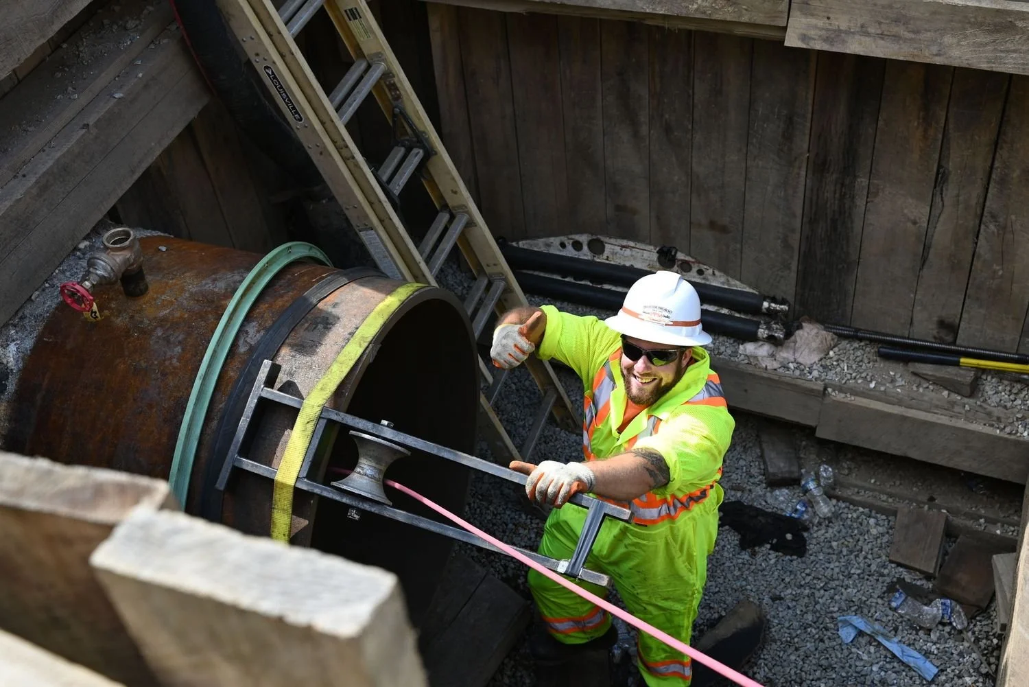 A construction worker in a neon green safety suit, white helmet, and sunglasses working on a large pipe at a construction site, smiling and giving a thumbs-up.