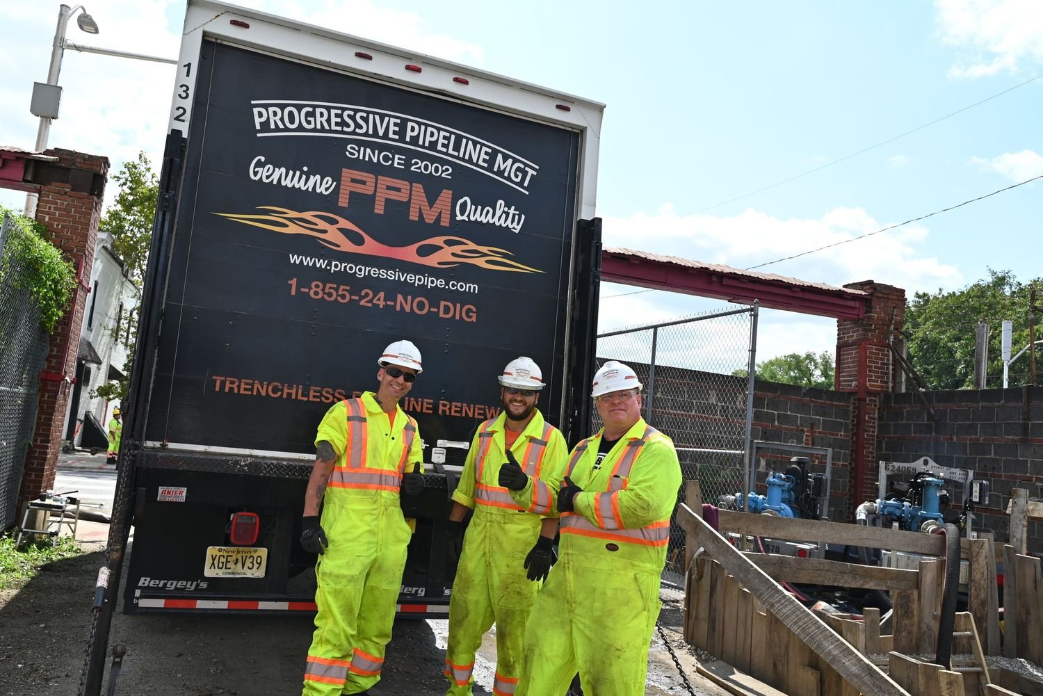 Three construction workers in bright yellow safety jumpsuits and white hard hats posing and smiling in front of a black trailer at a construction site. Two of them are giving thumbs up. The trailer has advertising for progressive pipe in white and re