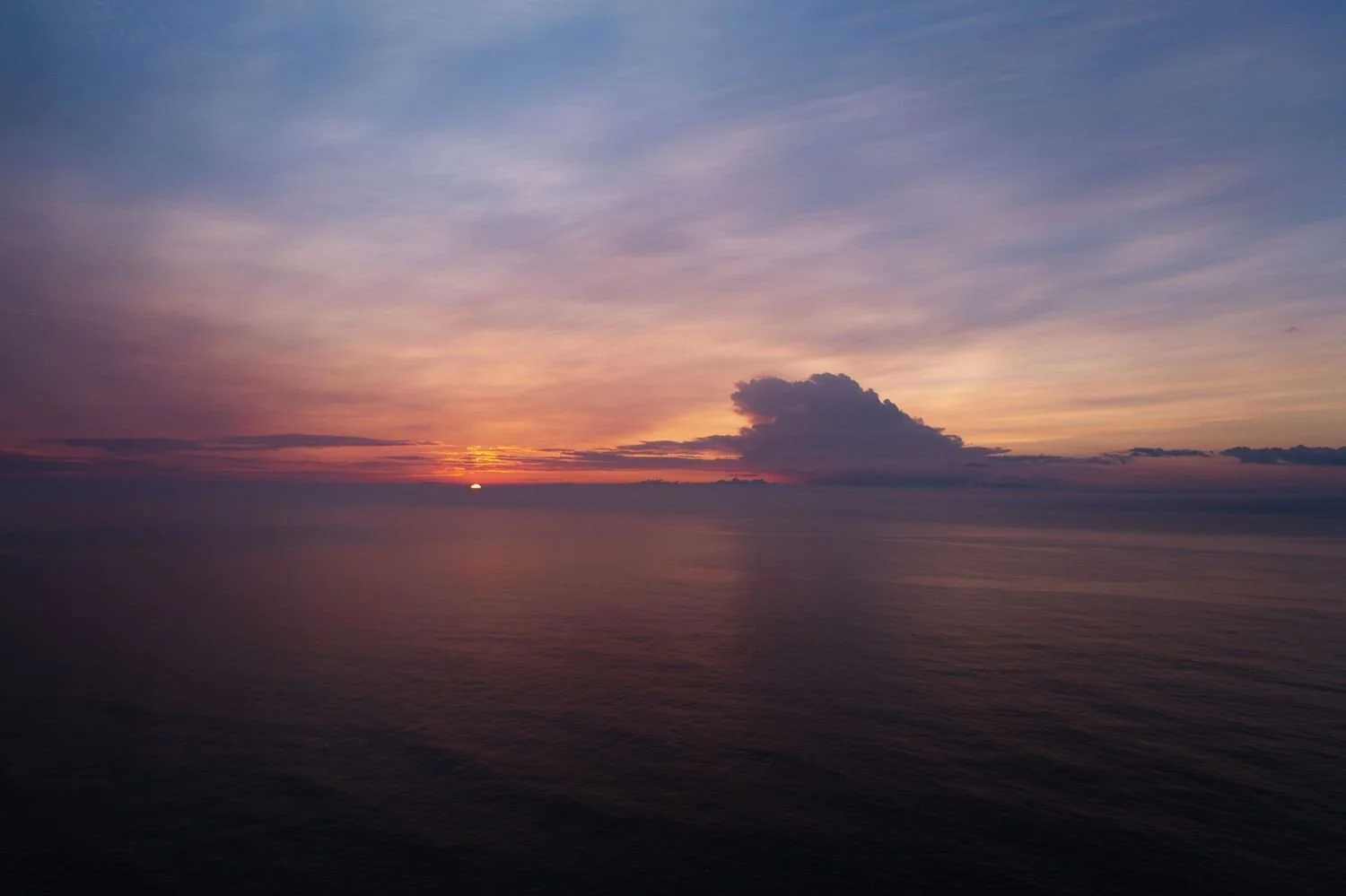 A sunset over a calm ocean with a partly cloudy sky, featuring warm hues of orange, pink, and purple.