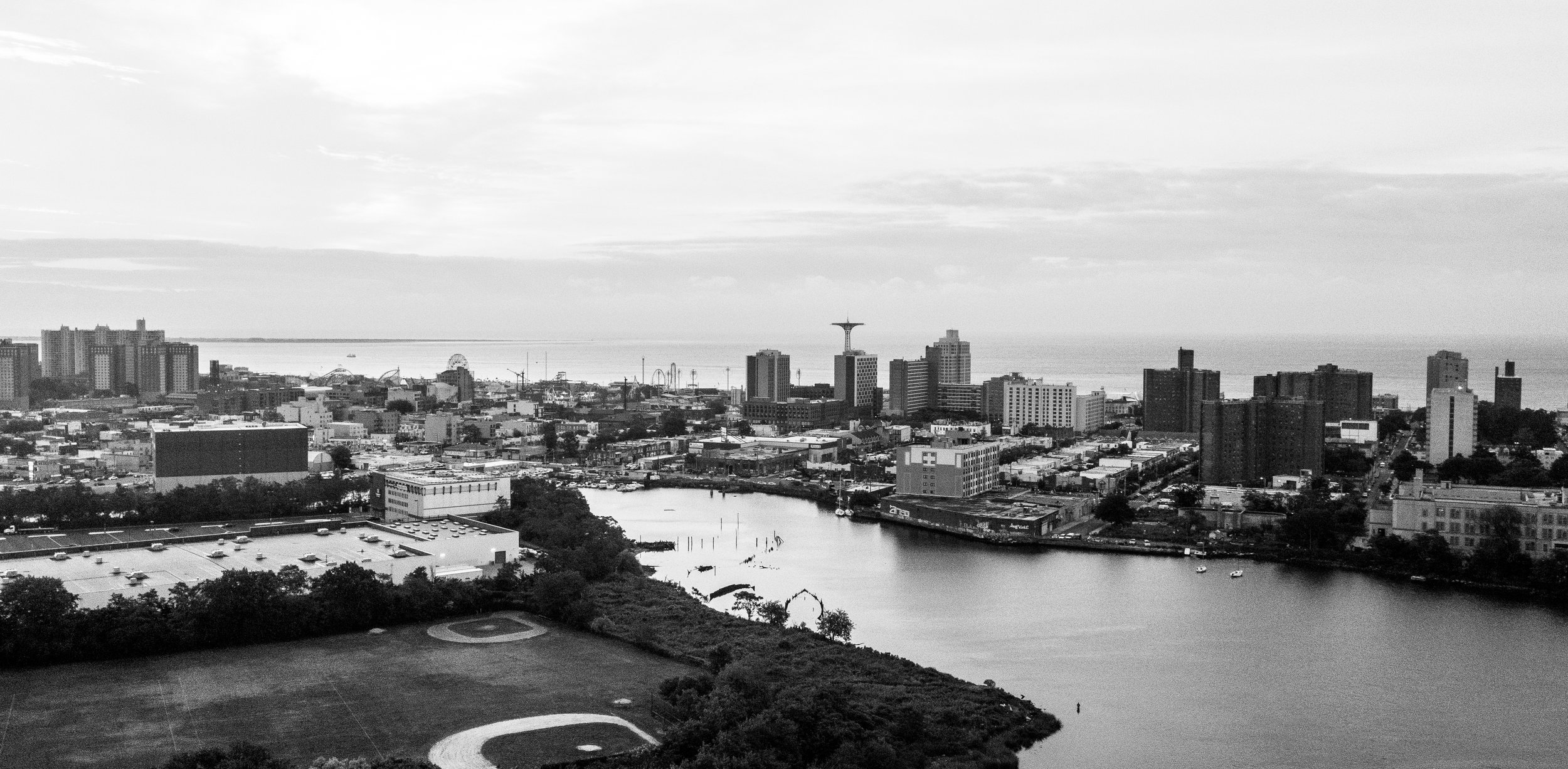 Black and white aerial view of a city with high-rise buildings, a harbor with boats, and a bridge, with a park and helipad in the foreground.
