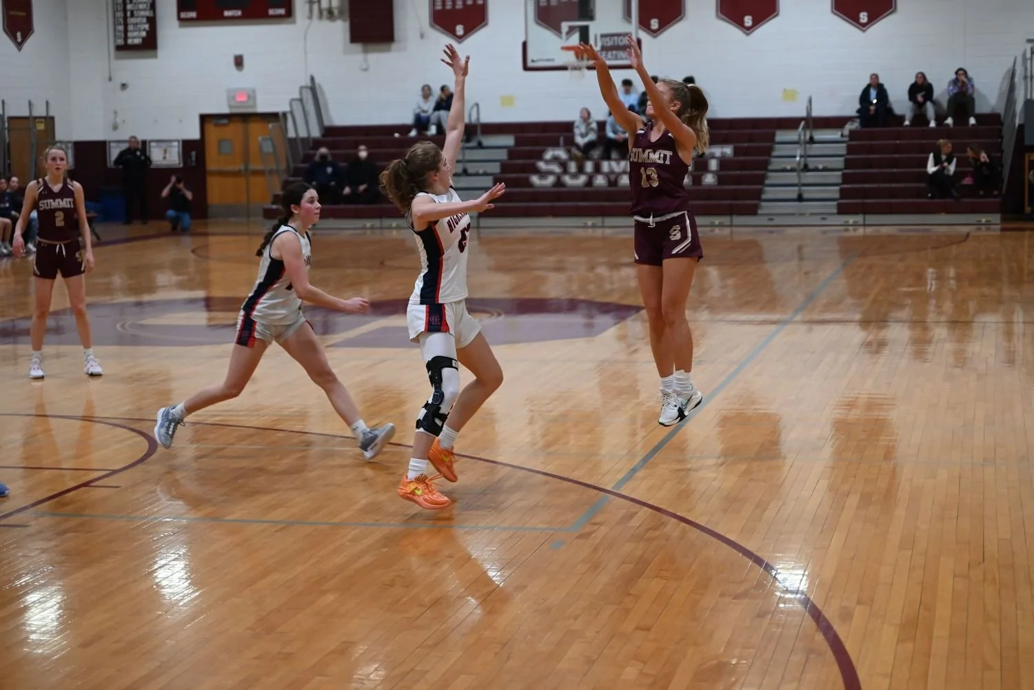 A girls' basketball game in progress on a gymnasium court with players in action, audience seated in the bleachers.