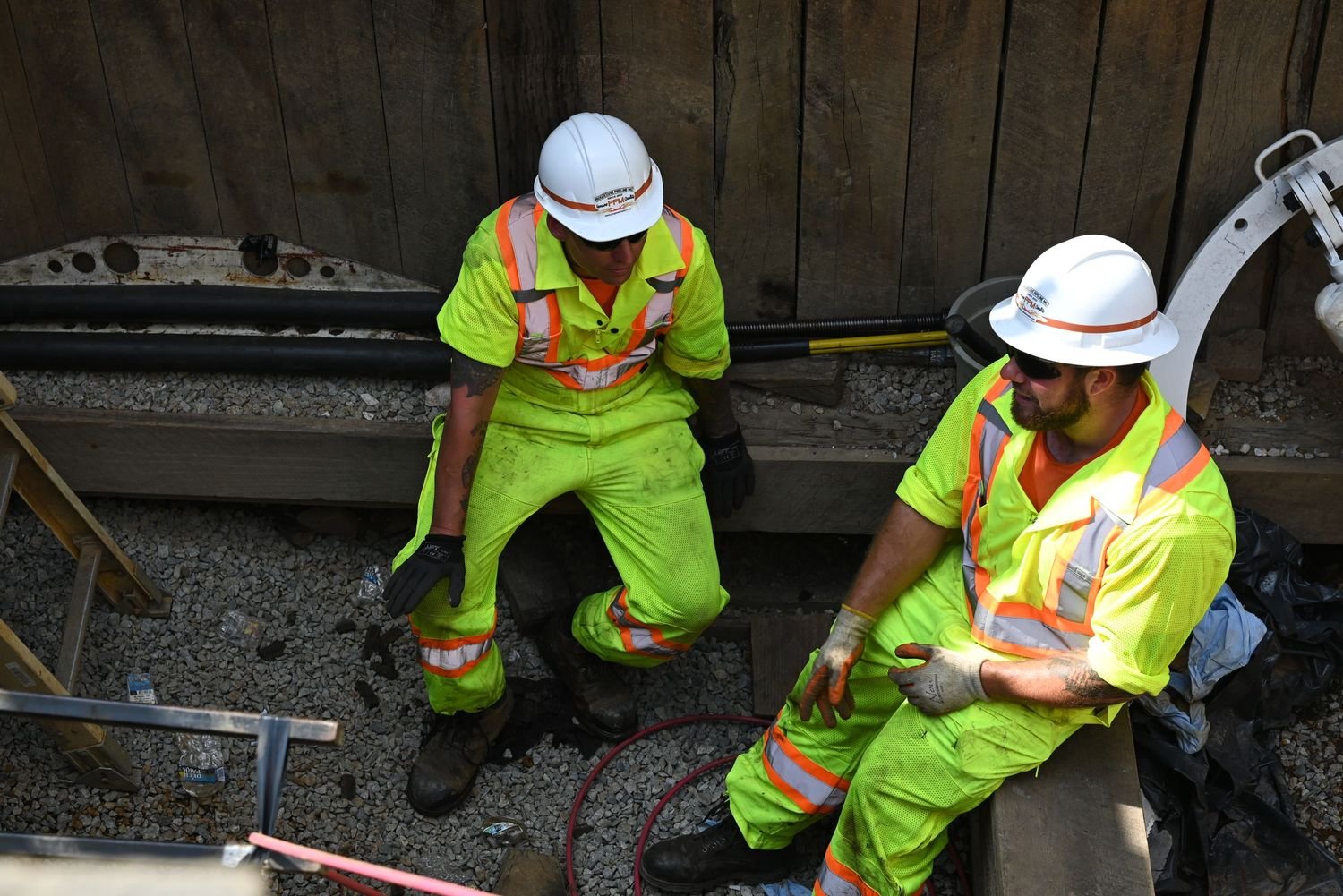 Two construction workers in neon yellow safety uniforms and white hard hats working on a construction site with gravel on the ground and wooden walls in the background.