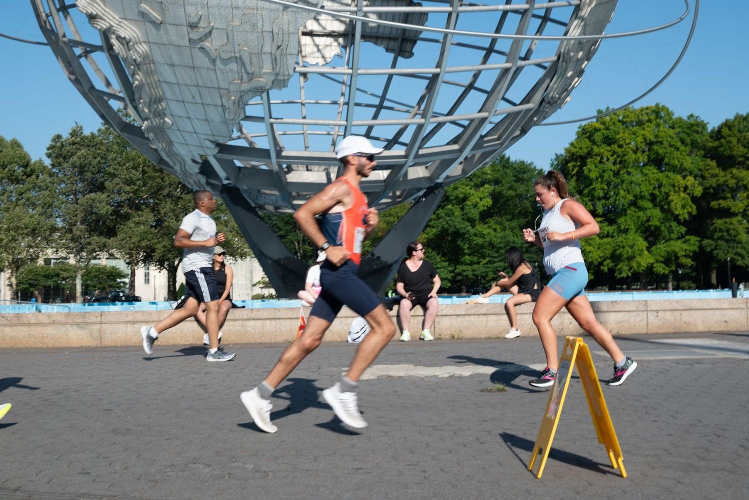 People participating in a marathon or running event near the Unisphere in a park with green trees, some sitting on a bench and others running.