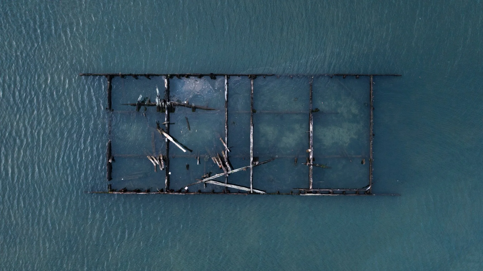 Aerial view of a rusted shipwreck on calm water, with its wooden and metal framework exposed and partially submerged.