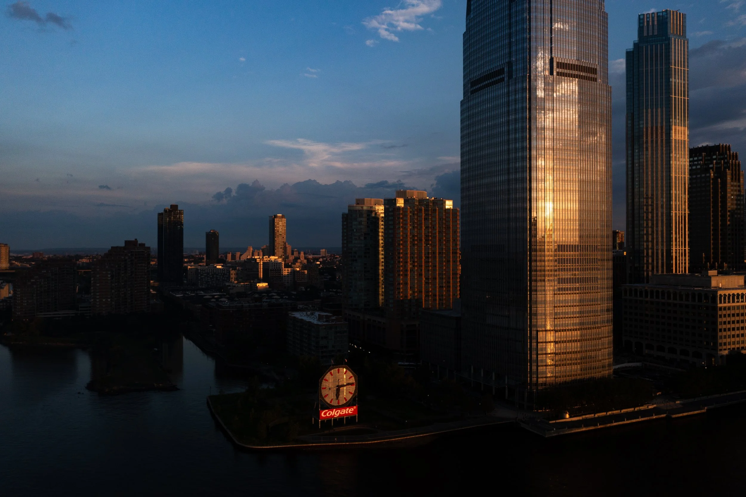 Skyline of tall modern glass skyscrapers at sunset with a river in the foreground and an advertisement sign for Colgate visible.