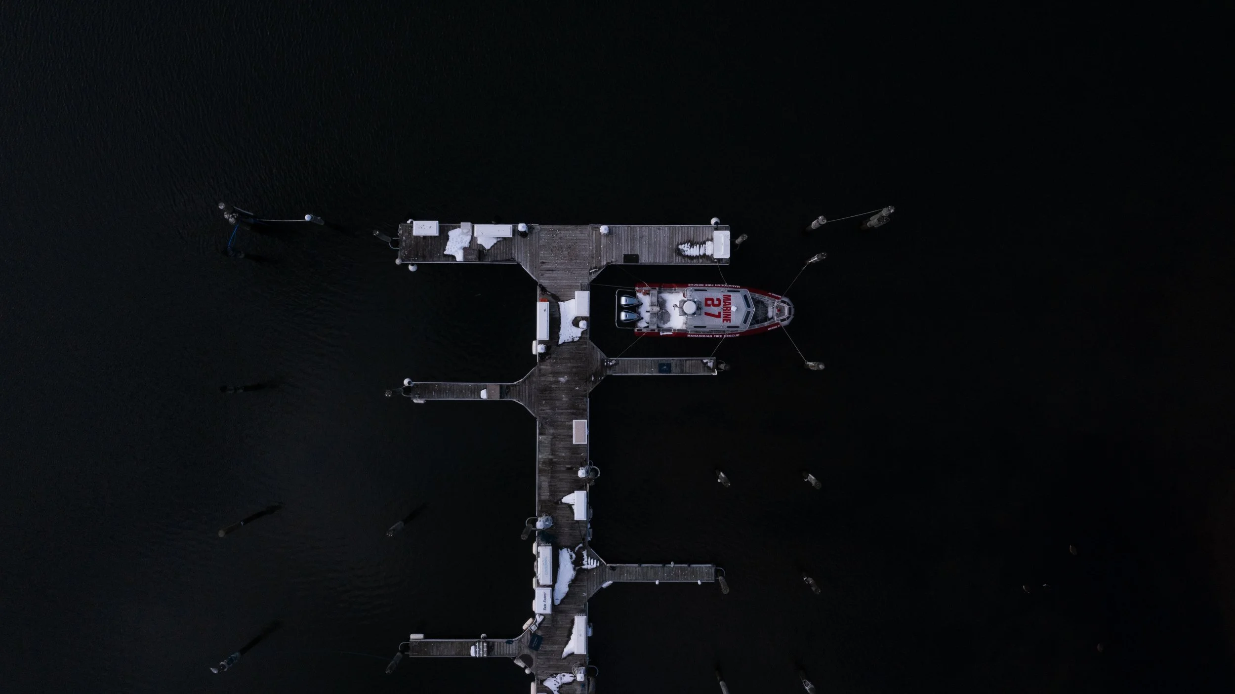 Aerial view of a wooden dock extending into dark water, with a red and white rescue boat tied to it, snow on the dock.