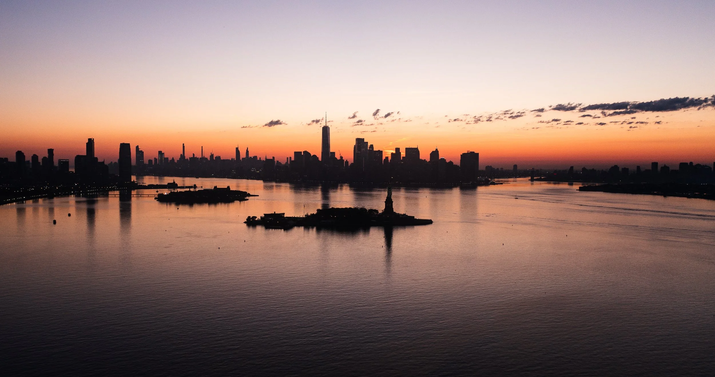 Silhouette city skyline at sunset over a river with Statue of Liberty in the foreground.