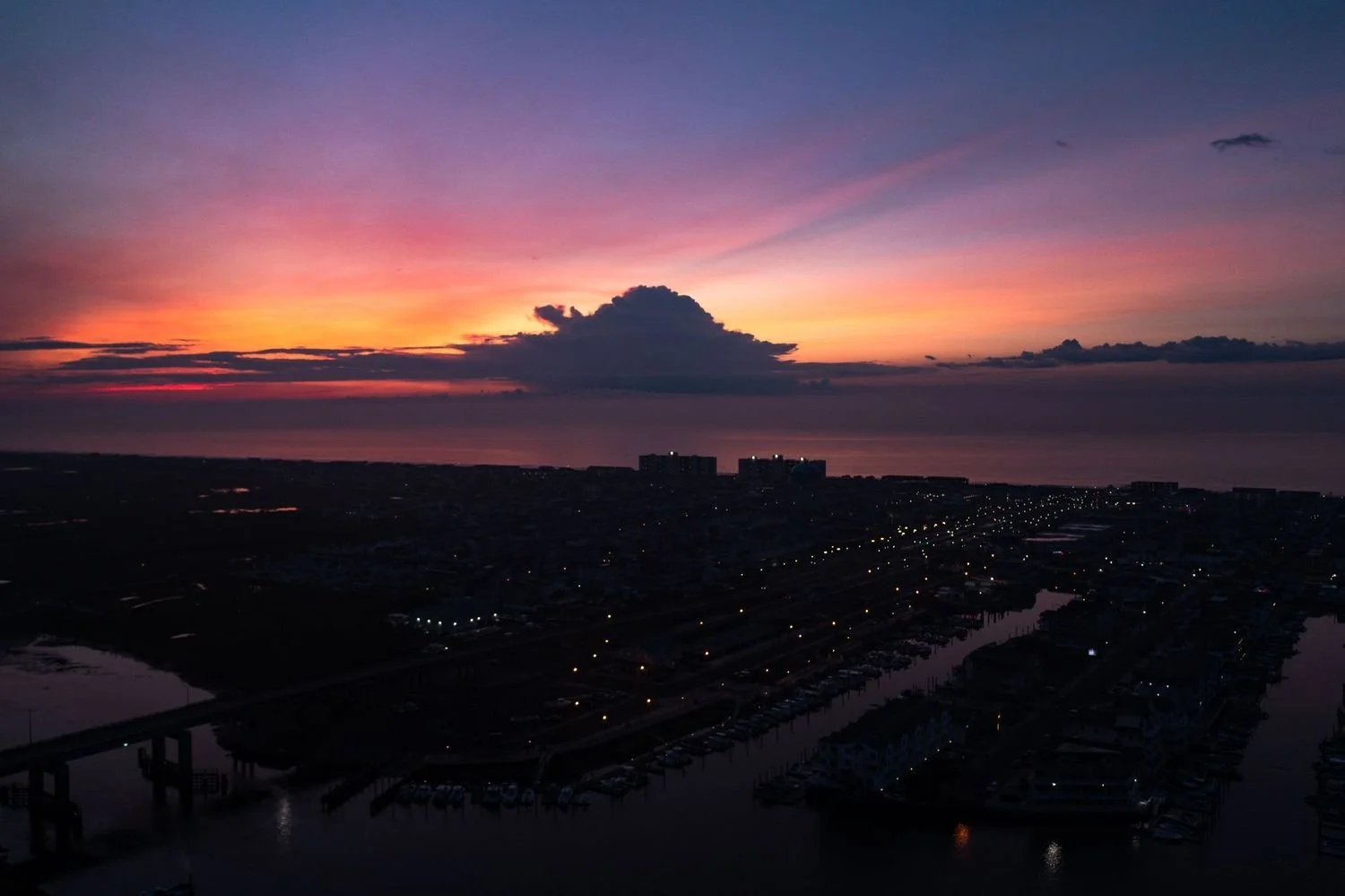 Sunset over a coastal city with colorful sky, harbor with boats, and city buildings in the distance.