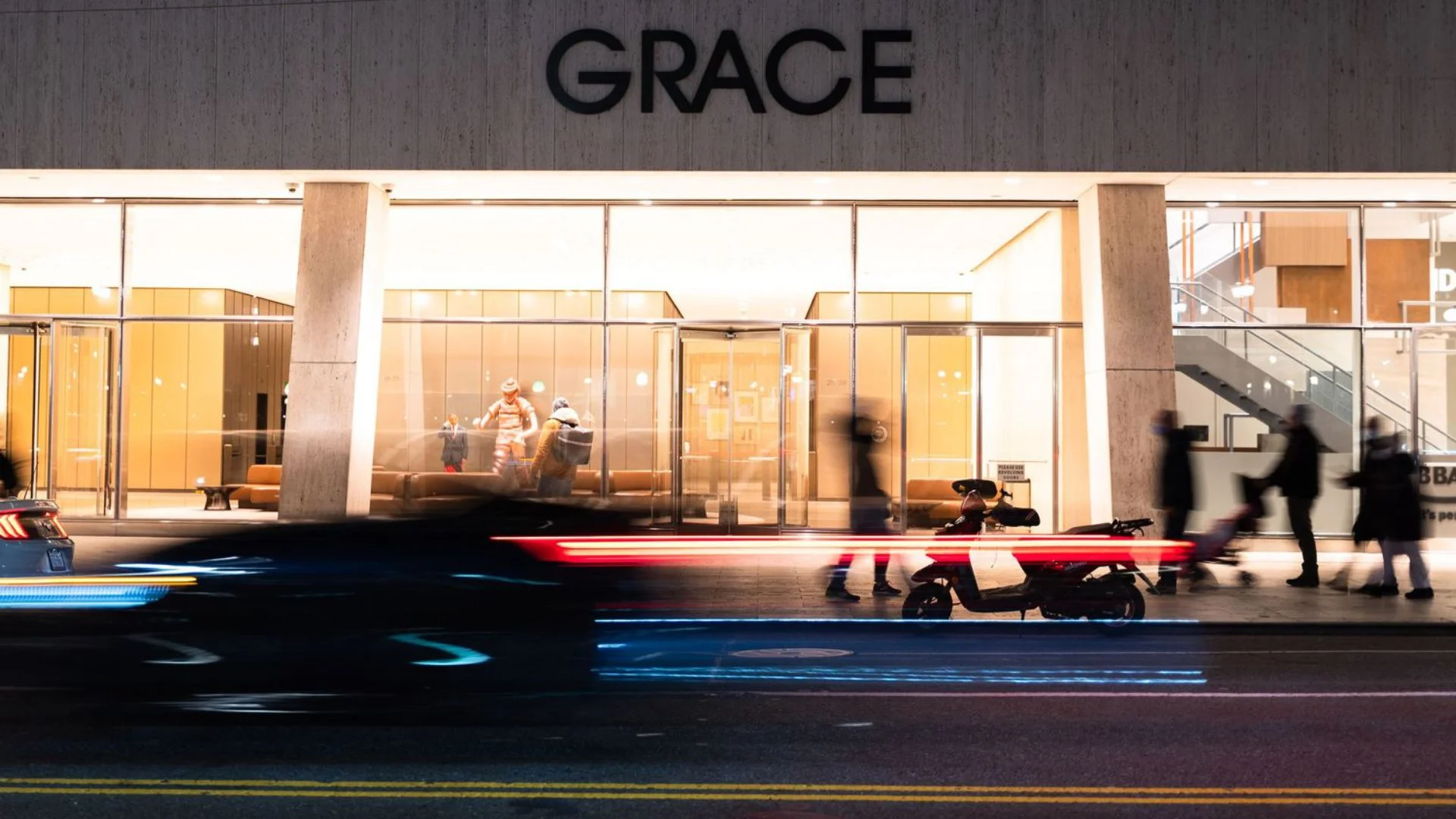 Night view of the Grace building with people walking and cycling in front, blurred car lights on the street, and some visible bikes near the sidewalk.