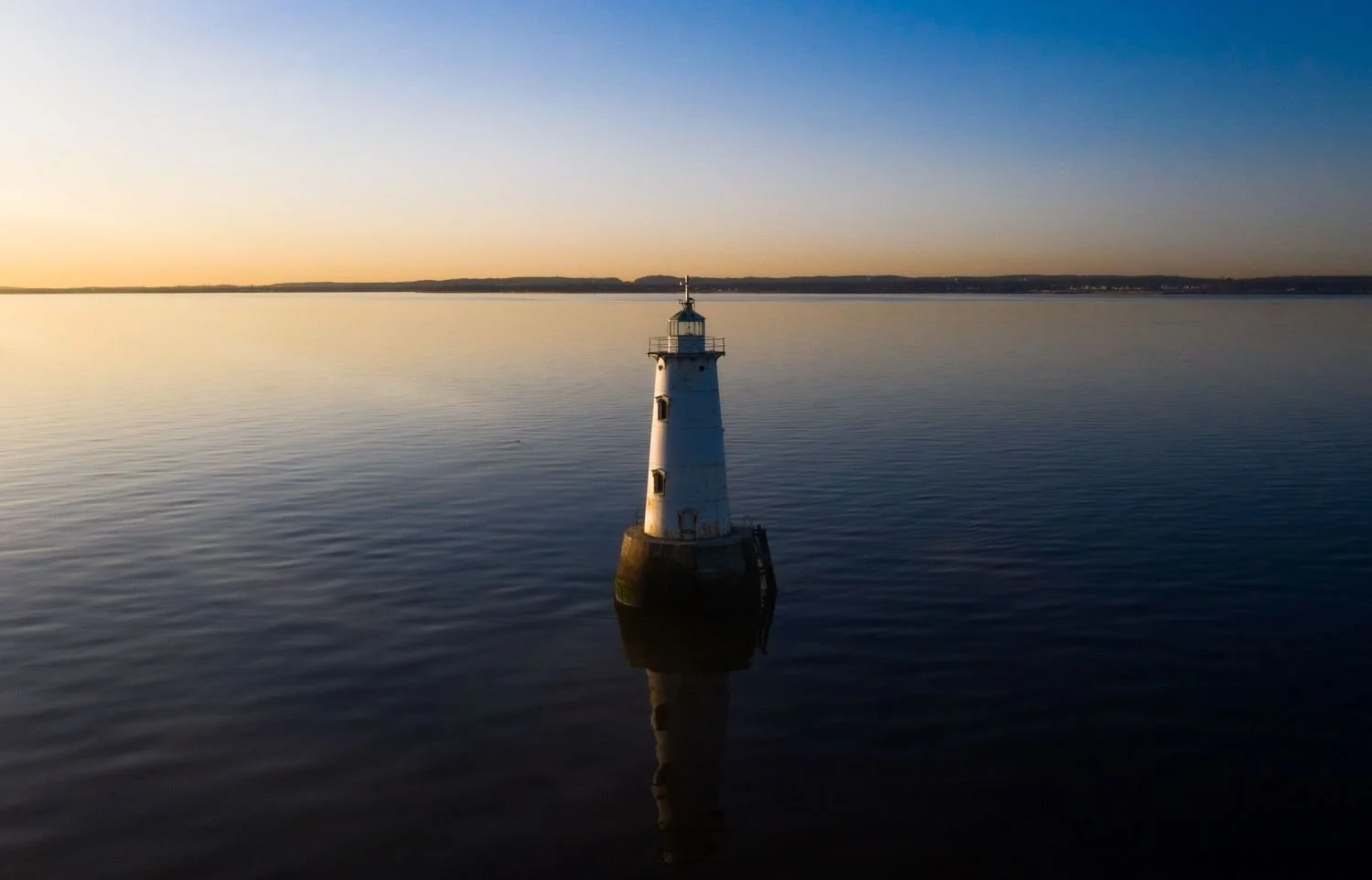 A lighthouse on the water at sunset or sunrise with a calm surface and a clear sky.