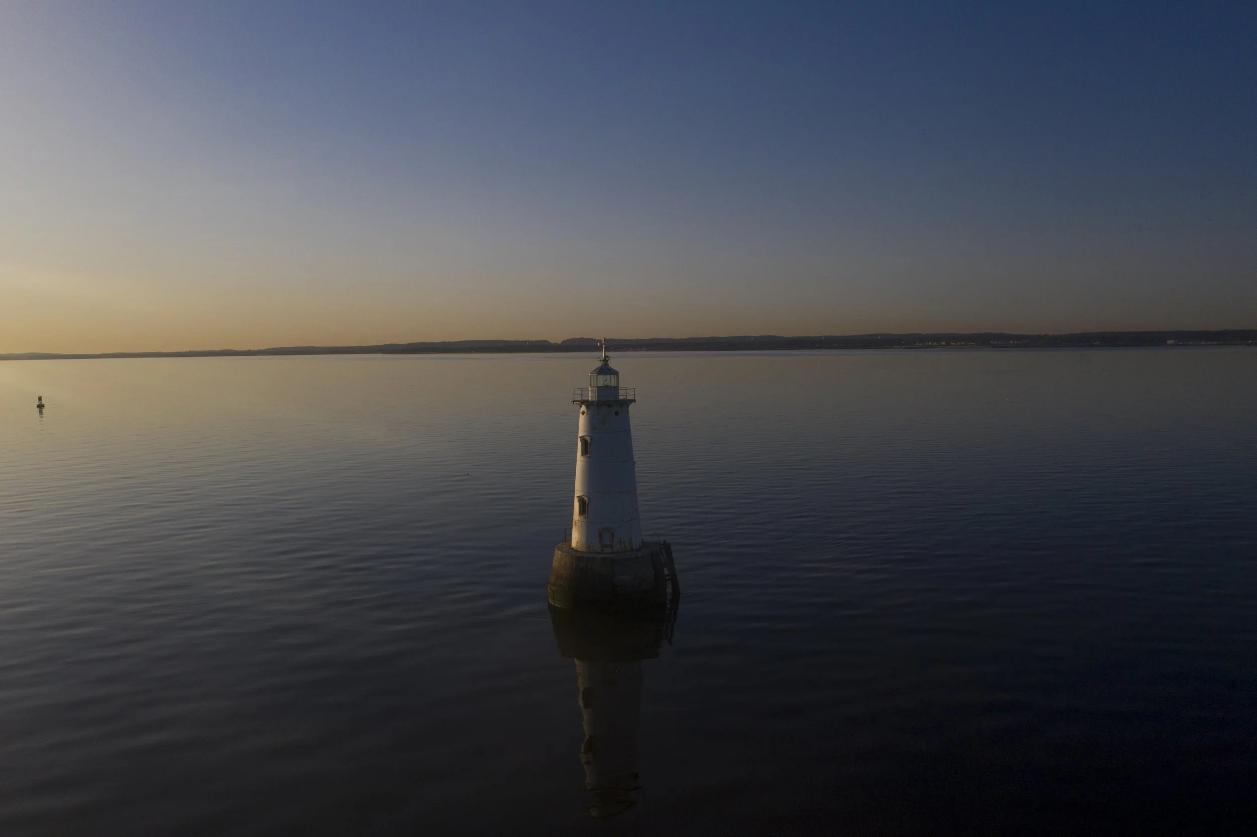 A lighthouse situated on a calm body of water during sunset with a clear sky and faint horizon line.