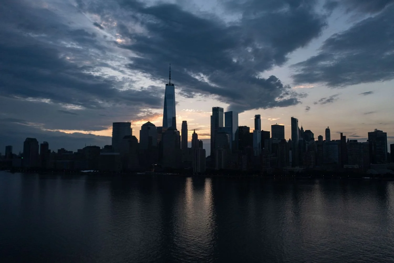 Sunset over New York City skyline, including One World Trade Center, with dark clouds in the sky and reflected in the water.