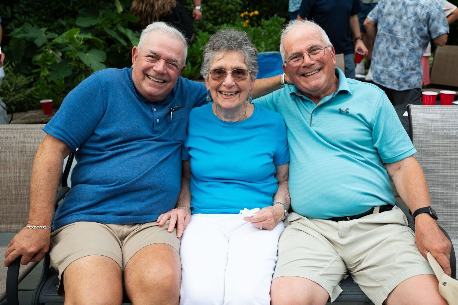 Three smiling older adults, two men and one woman, sitting closely together outside, at a gathering or party, with other people and red cups visible in the background.