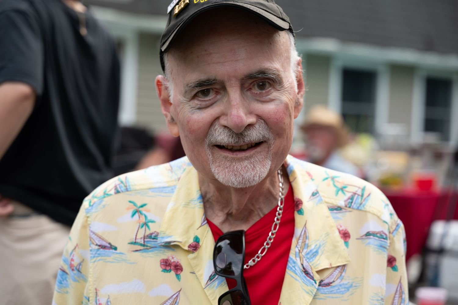 An elderly man with a white beard and mustache, wearing a tropical print yellow Hawaiian shirt, red t-shirt, sunglasses hanging from his shirt collar, and a silver chain necklace, smiling at an outdoor gathering.