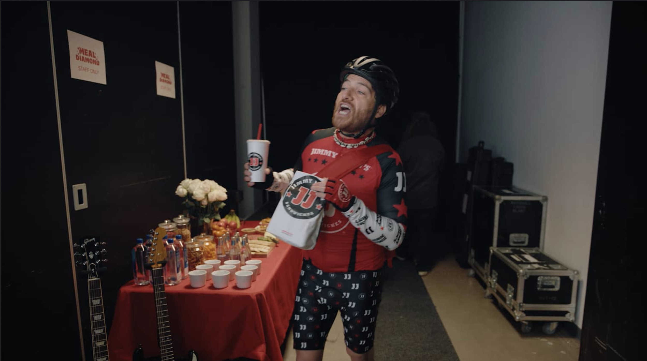 A man wearing a red cycling jersey, black helmets, and pajamas with a J.J. logo, standing at a table with food and cups, holding a drink and a snack bag, in a backstage or event area with musical instruments and equipment.