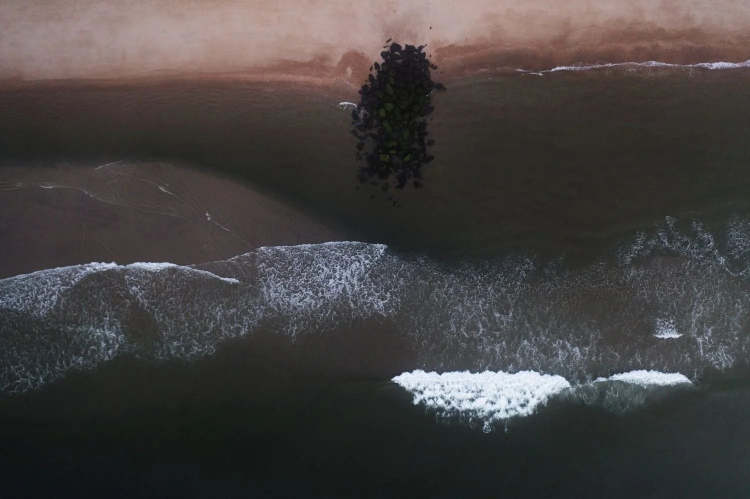 Aerial view of waves crashing onto a sandy beach with rocks and a jetty extending into the ocean.