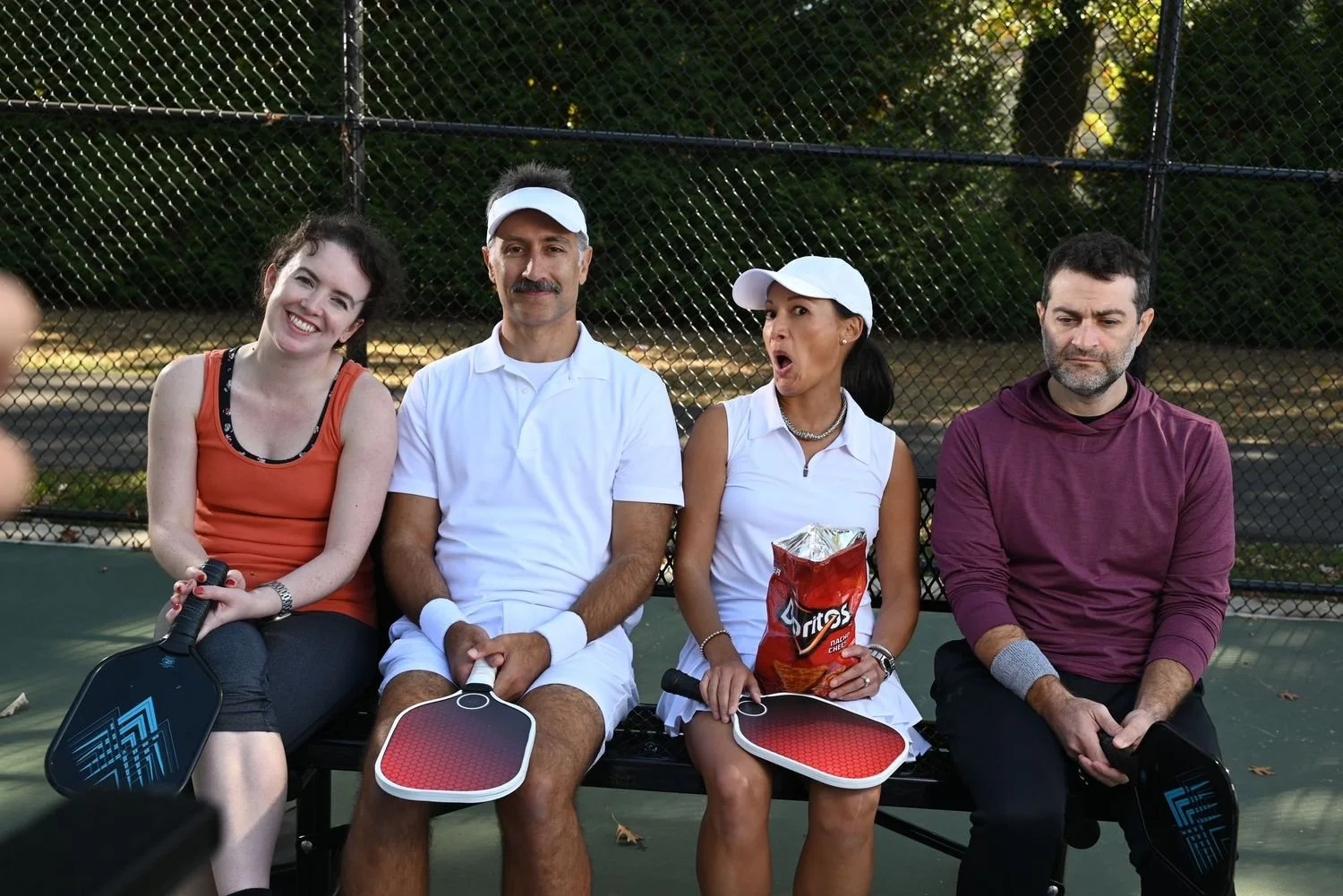 Four people sitting on a bench at a tennis court, holding paddles, with a chain-link fence and trees in the background. The group includes a smiling woman in an orange tank top, a man with a mustache in a white polo shirt, a woman in a white shirt wi