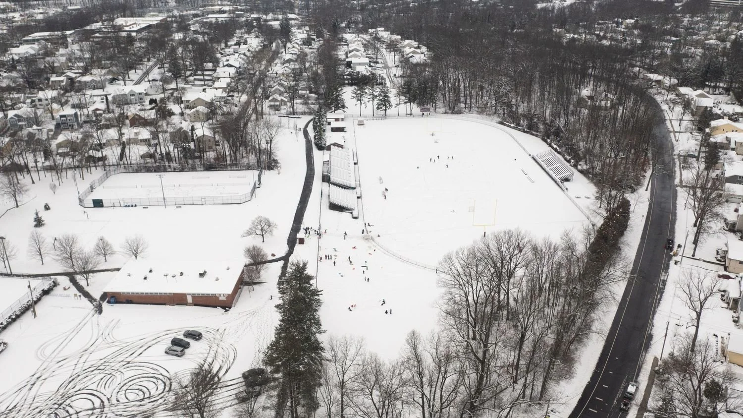 Aerial view of a snow-covered park with a skating rink, a baseball field, and a large grassy area. Surrounding residential houses and streets are also visible.