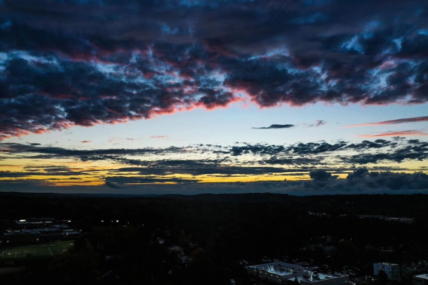 A sunset sky with dark clouds and a mix of yellow, pink, and blue hues, overlooking a town with buildings and trees in the foreground.