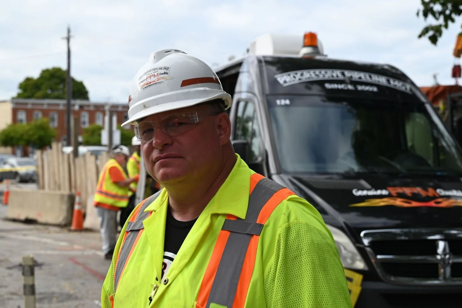 A construction worker in a safety vest, hard hat, and safety glasses stands in front of a work truck on a construction site, with other workers and orange traffic cones in the background.