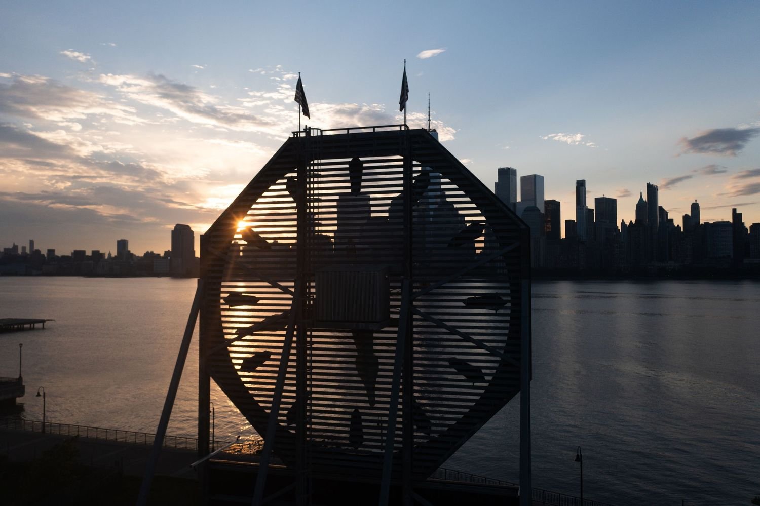 A clock installation with a nautical theme silhouetted against a sunset skyline over a body of water, with a city skyline in the background.