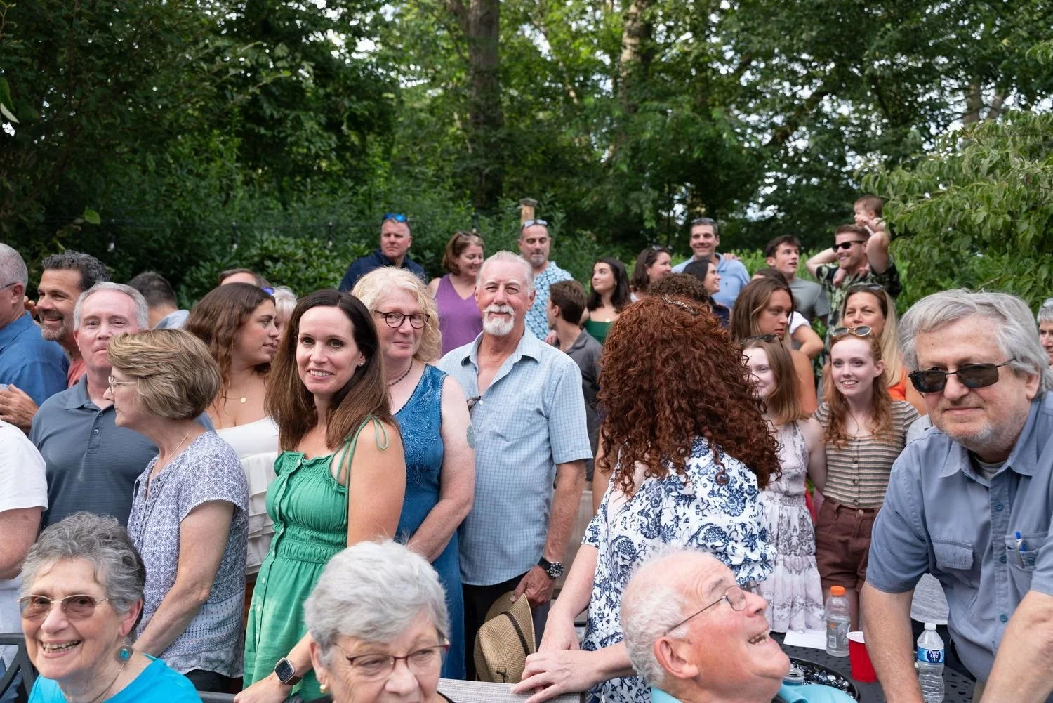 A large crowd of people gathered outdoors, some smiling and talking, with trees in the background, at what appears to be a social event or gathering.