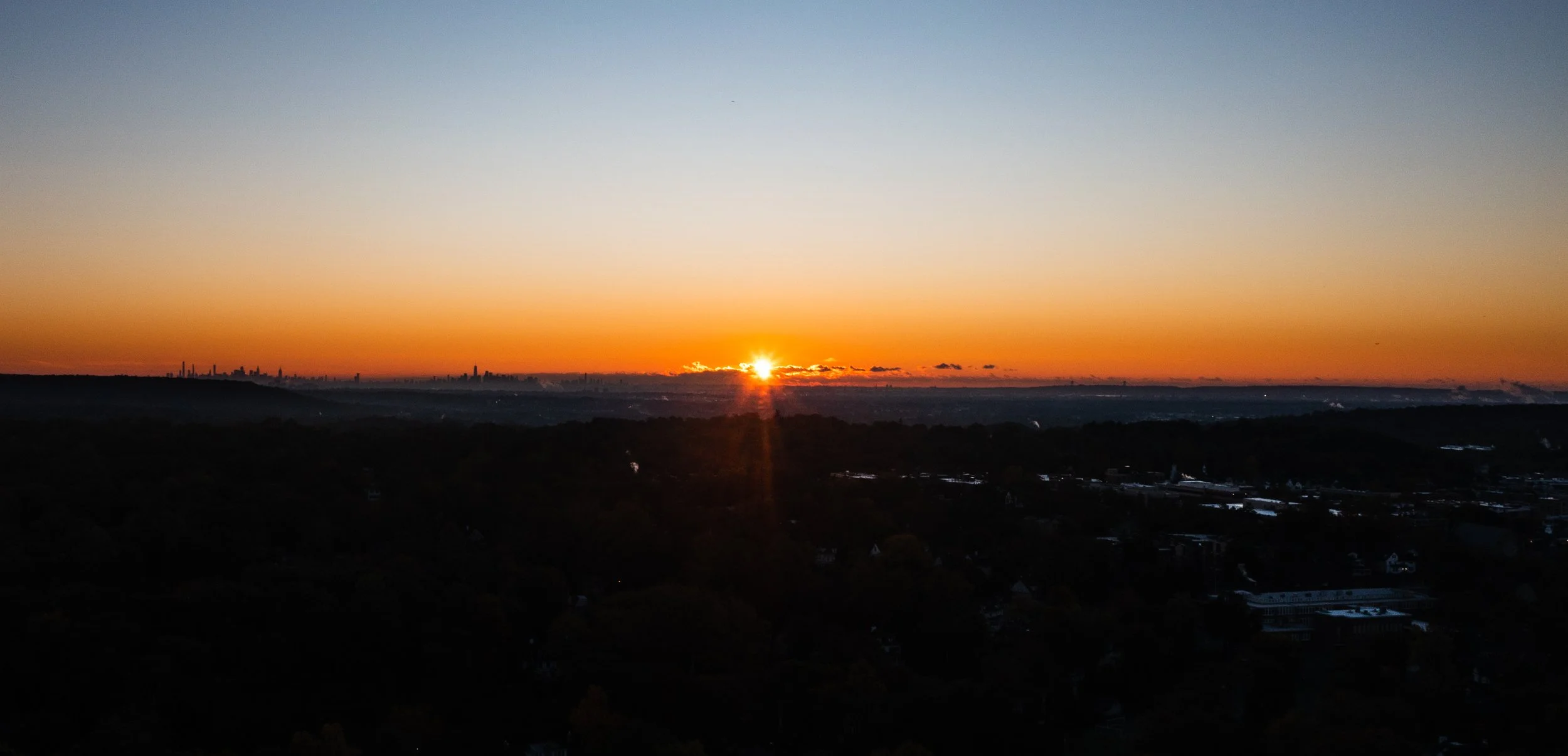 Sunset over a city skyline with orange and yellow hues in the sky, partly cloudy, with a silhouette of trees and buildings in the foreground.