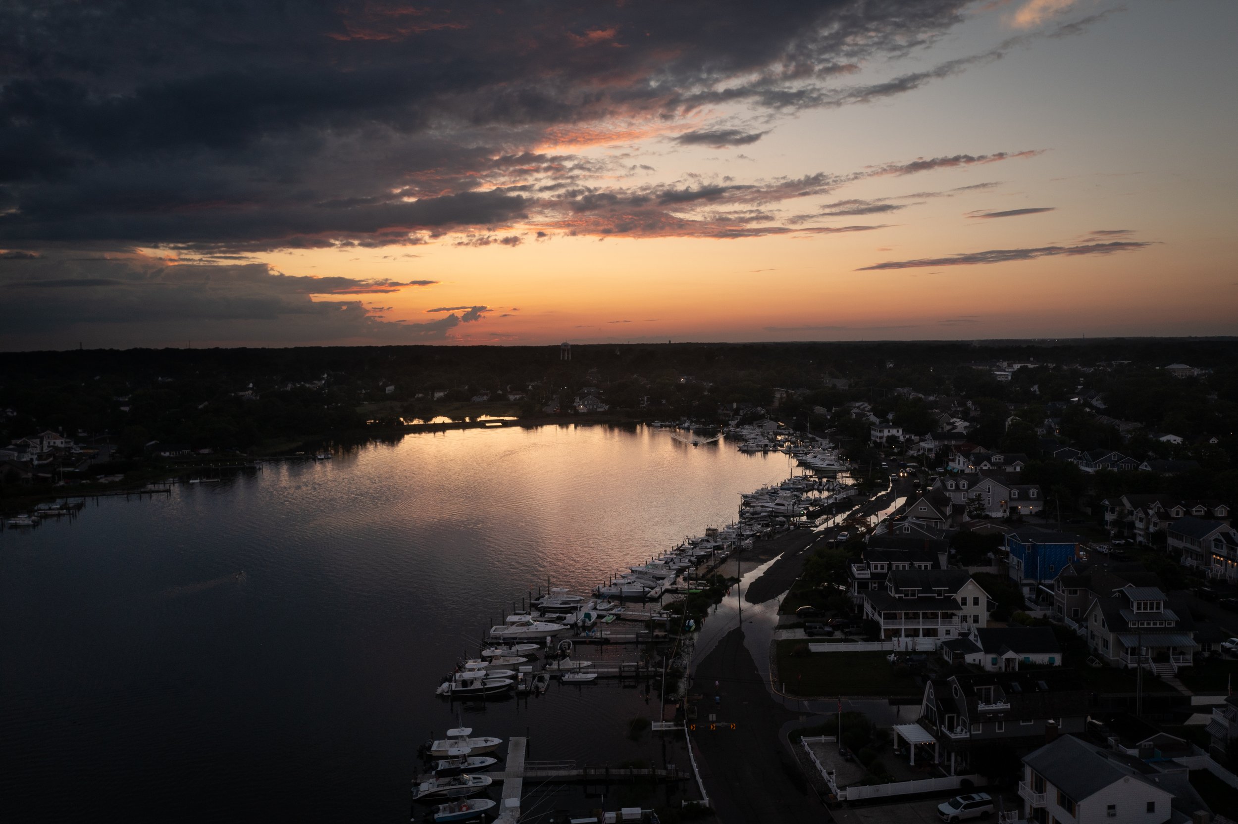 Sunset over a residential harbor with boats docked along the water, houses lining the shore, and dark clouds in the sky.