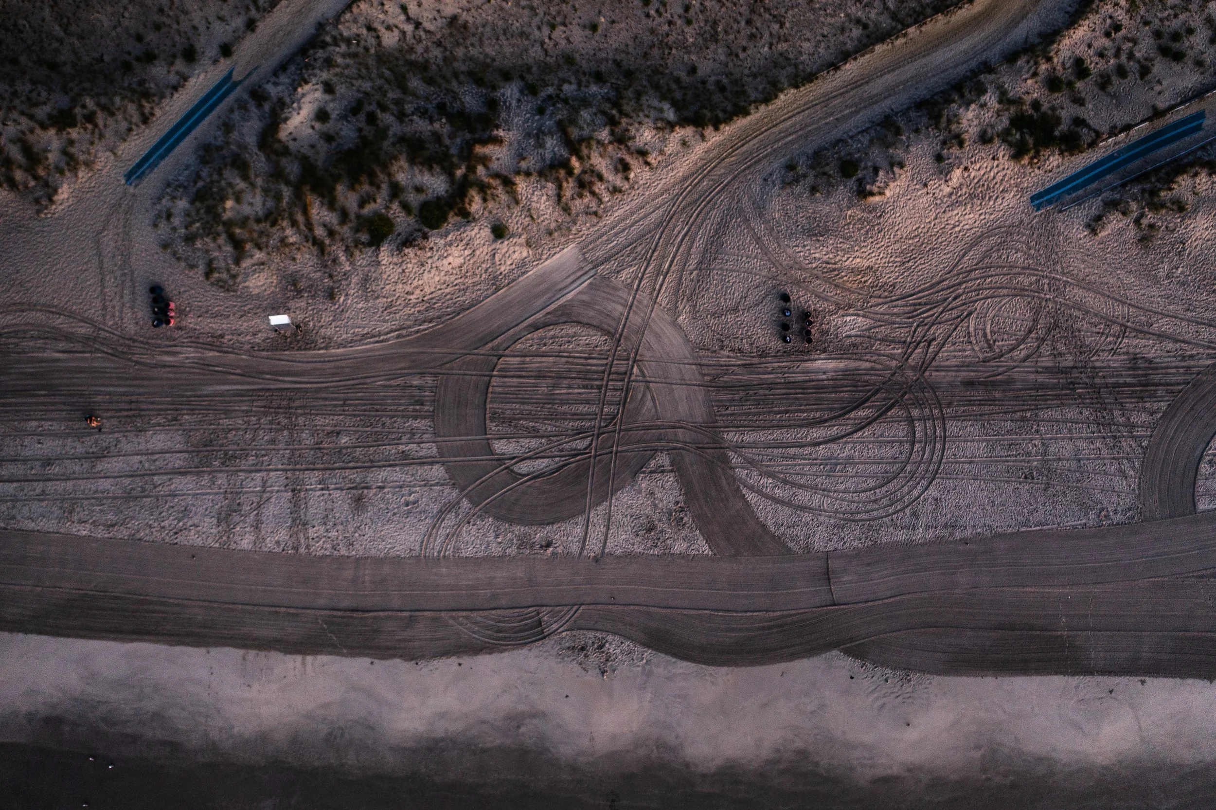An aerial view of a dirt area with multiple tire tracks and two blue long structures on opposite sides, surrounded by sparse bushes and a few cars.