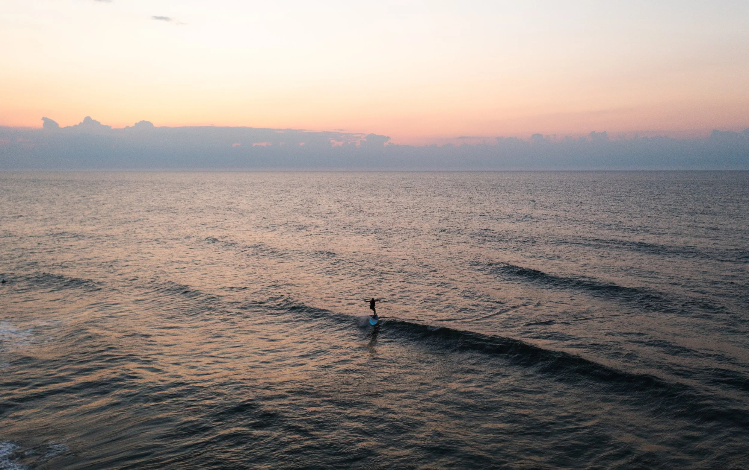 A person riding a paddleboard on the ocean during sunset, with a sky filled with soft pastel colors and a few clouds.