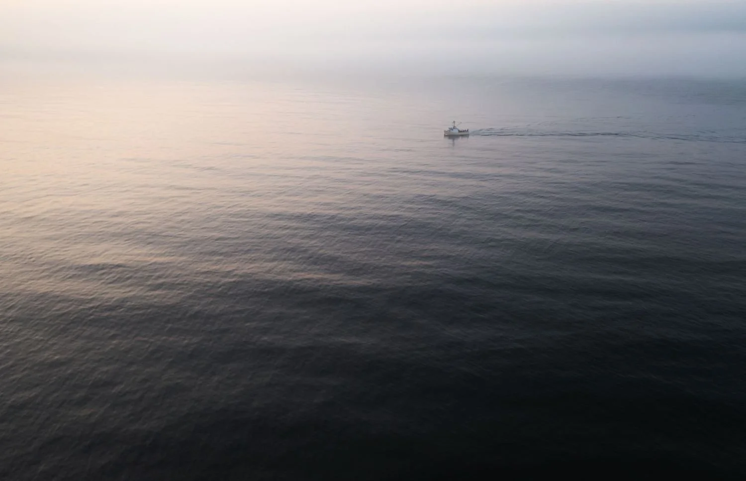 A small boat sailing across calm, dark ocean waters during dusk, leaving a gentle wake behind.