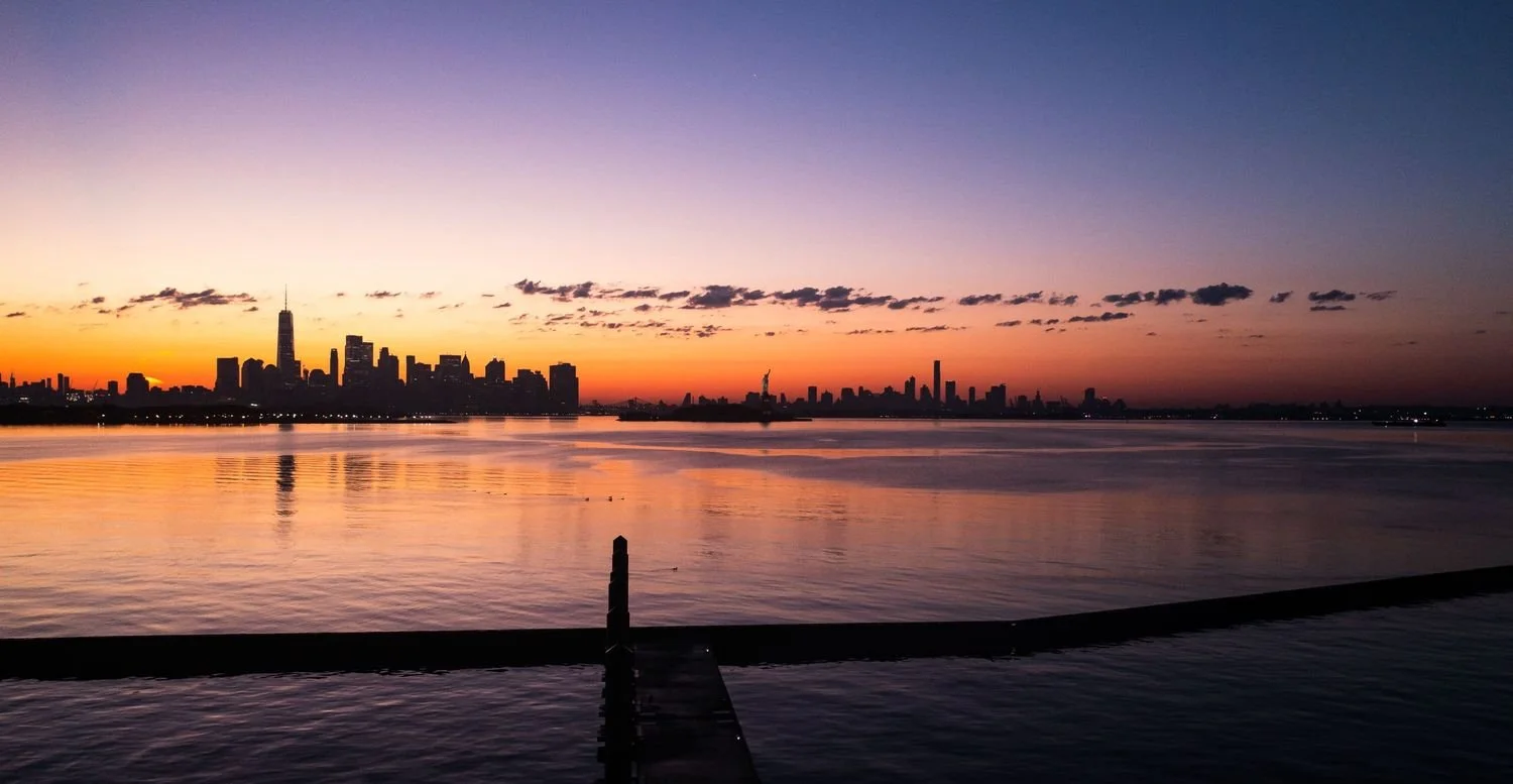 City skyline at sunset with a body of water in the foreground and a dock extending into the water.