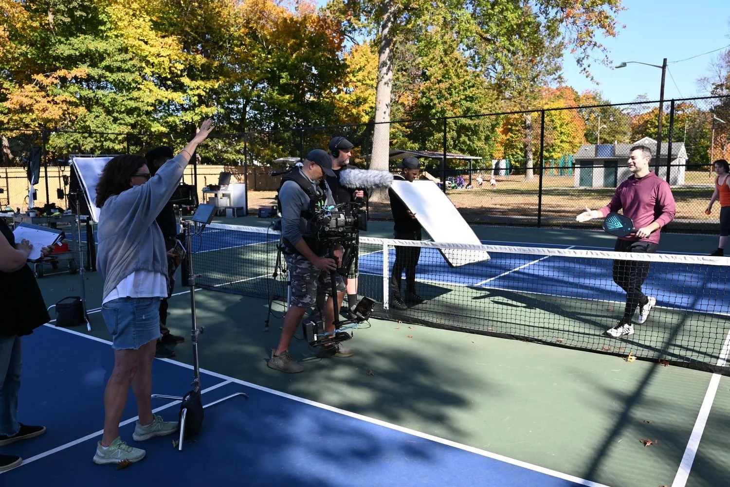 Film crew filming a man on a tennis court with a boom mic, while she mentions to camera.