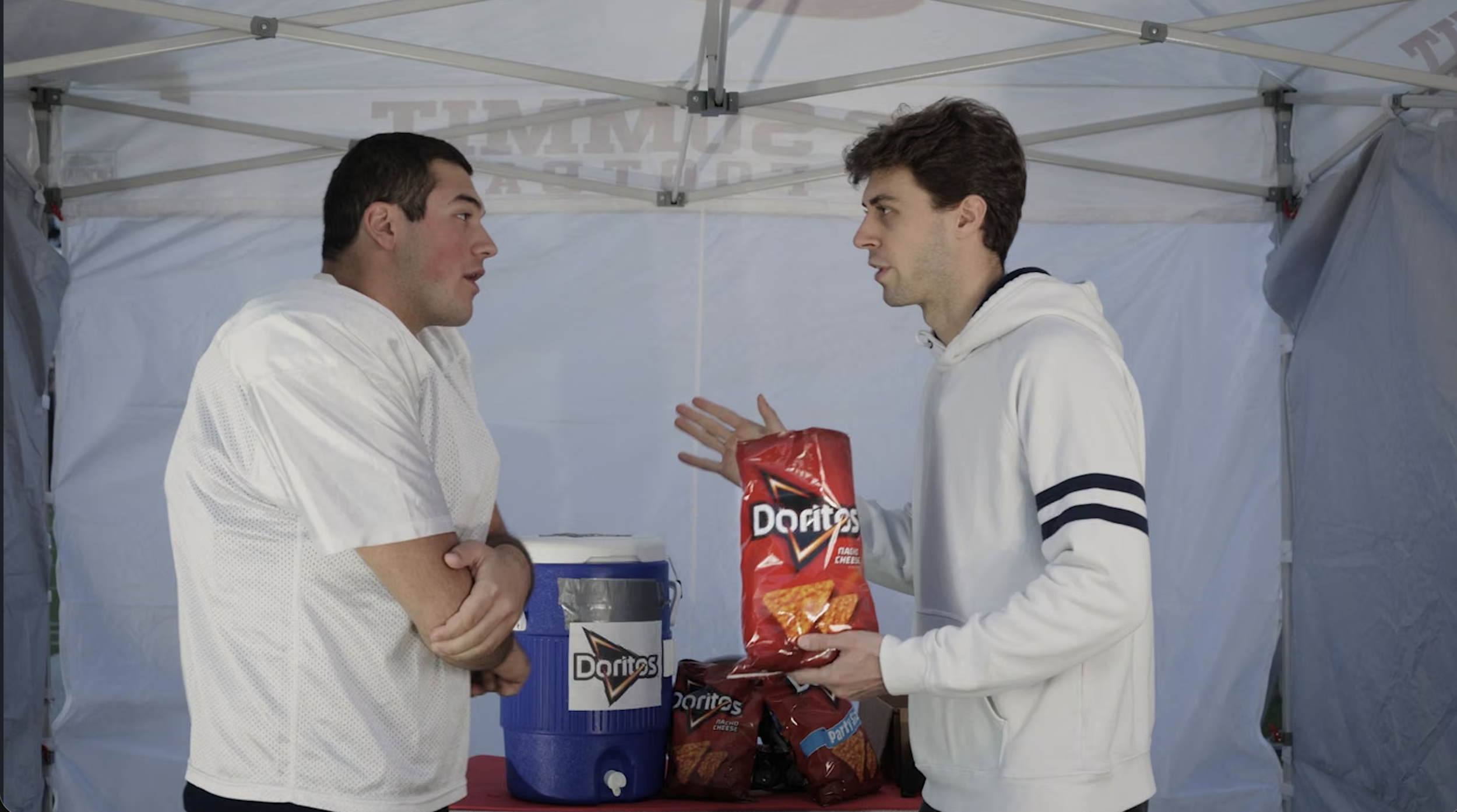 Two young men with light skin inside a white canopy tent, holding a conversation. One is wearing a white football jersey and the other a white hoodie with black stripes. The man in the hoodie is holding a bag of Doritos chips, and there is a blue cooler and a large trash bin with a Doritos cup in the scene.