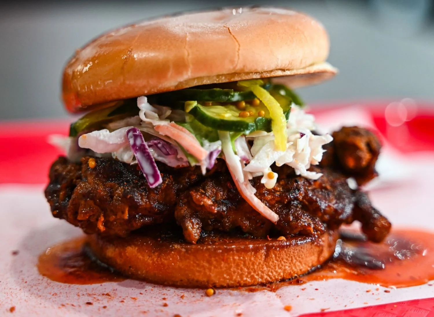 Close-up of a burger with a fried chicken fillet, coleslaw, pickles, and a toasted bun, with sauce drizzled around on a red and white surface.