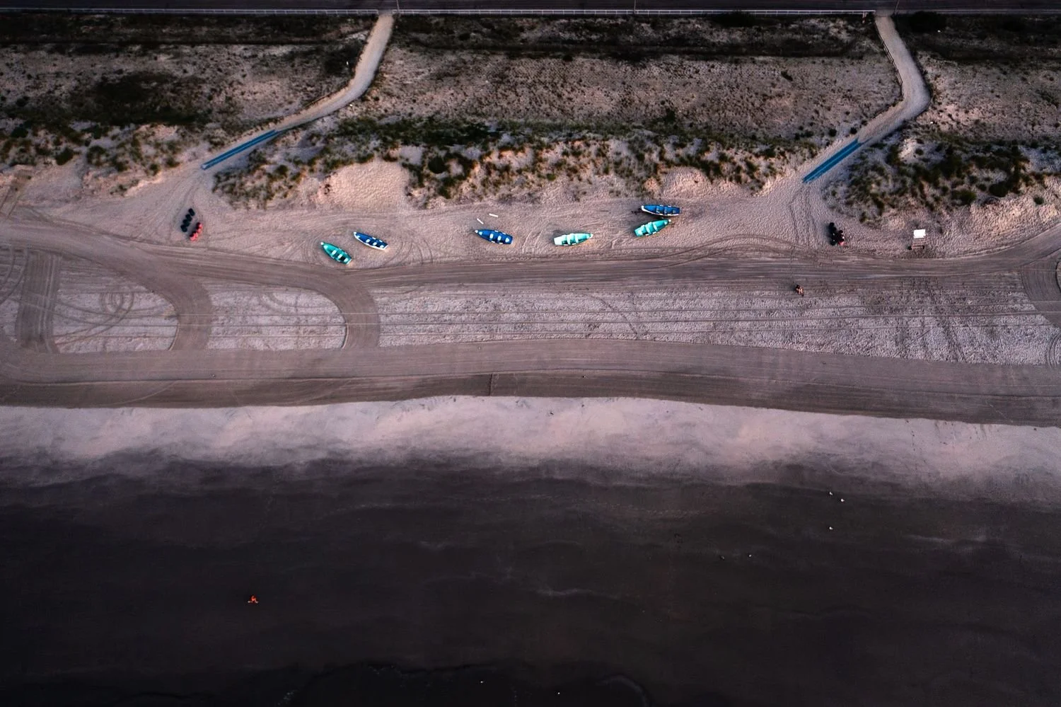 An aerial view of a sandy beach with colorful kayaks and paddleboards lined up, adjacent to a dark water shoreline, with some vehicles and structures in the background.