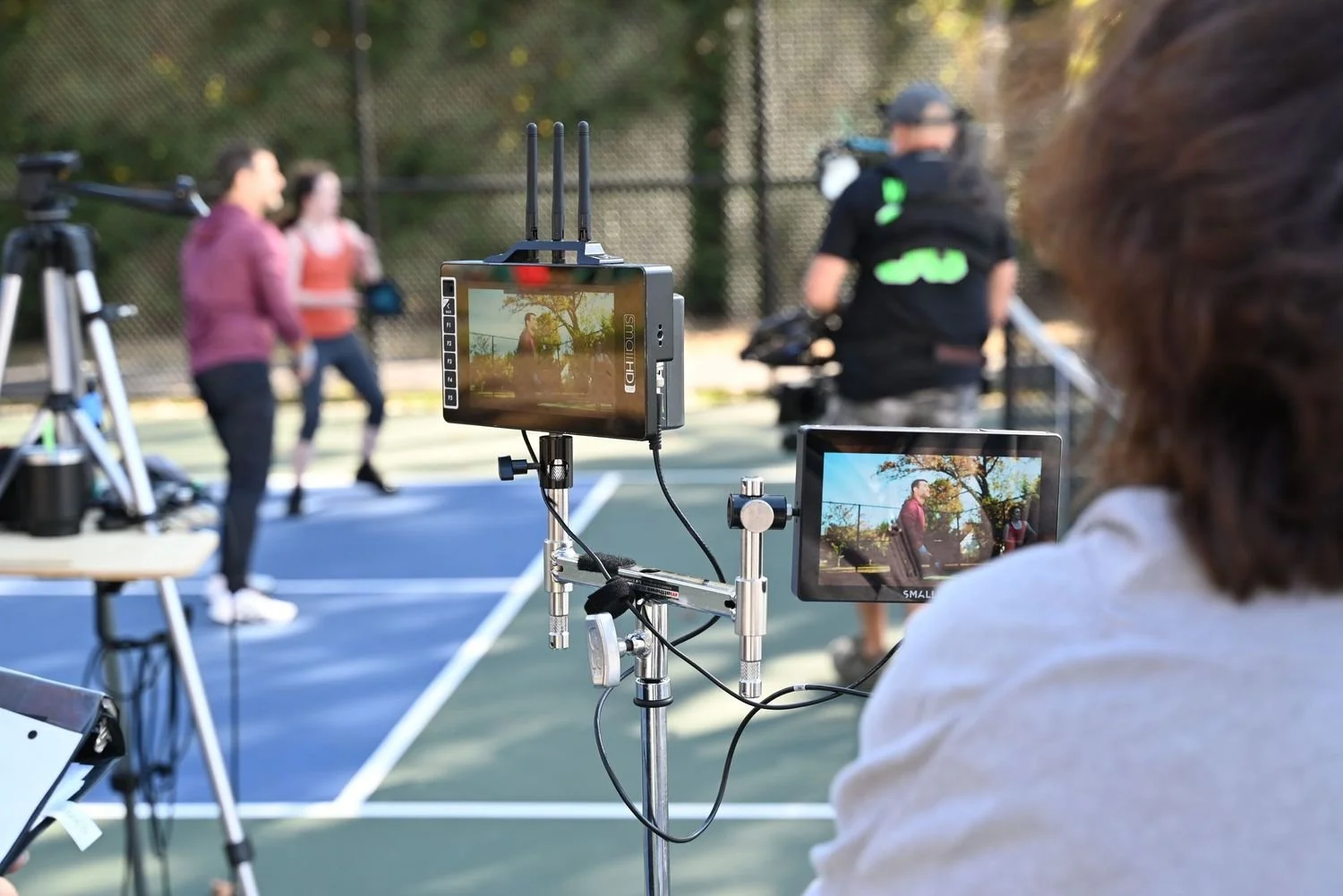 A film crew records a scene on a tennis court, with a camera crew, monitors, and people filming and acting.