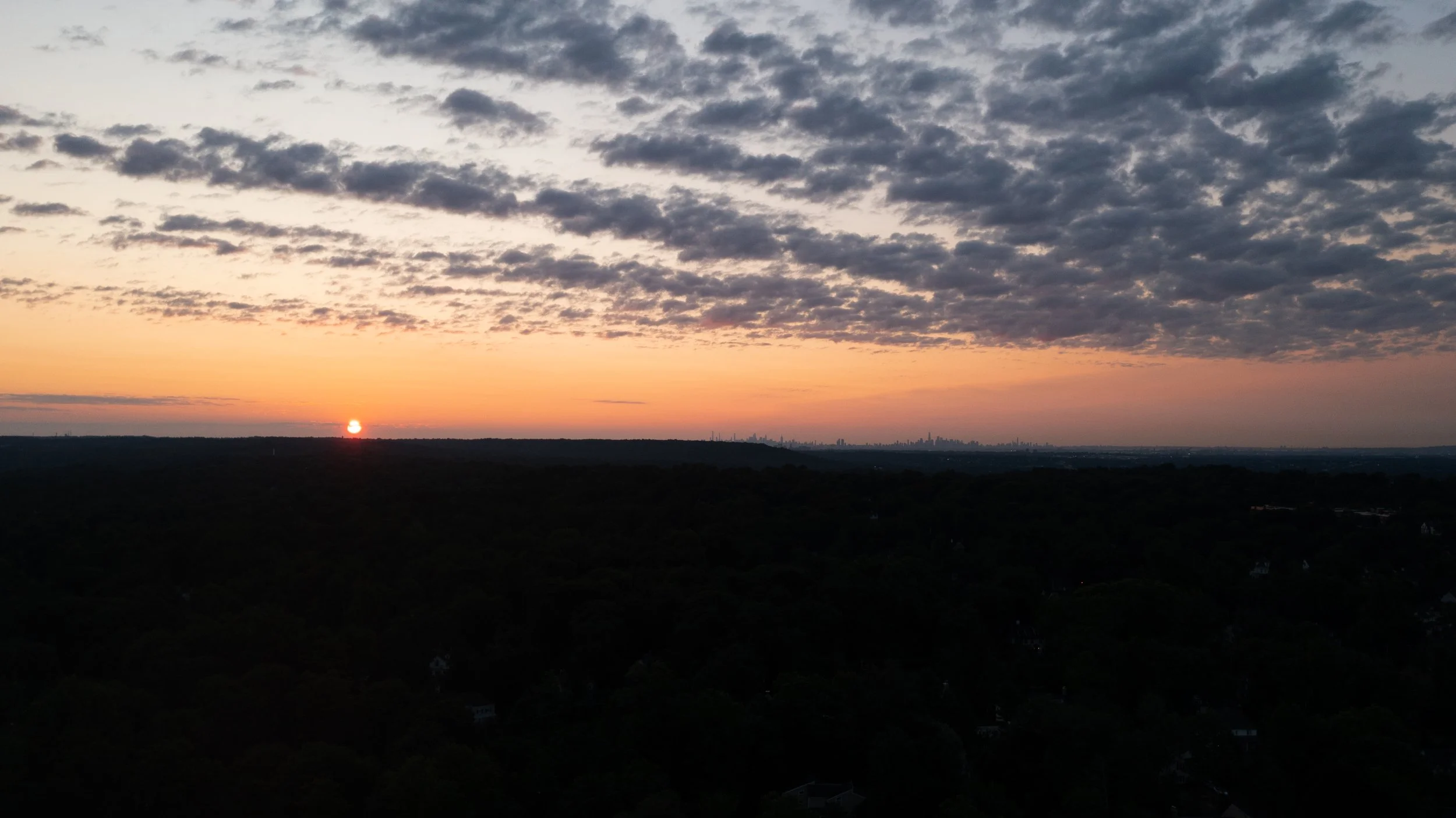 Sunset over a distant city skyline with scattered clouds in the sky.