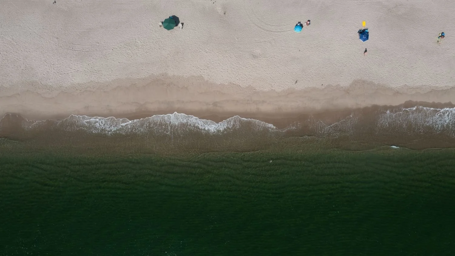An aerial view of a sandy beach with colorful umbrellas and people relaxing, with waves coming ashore and green water visible at the bottom.