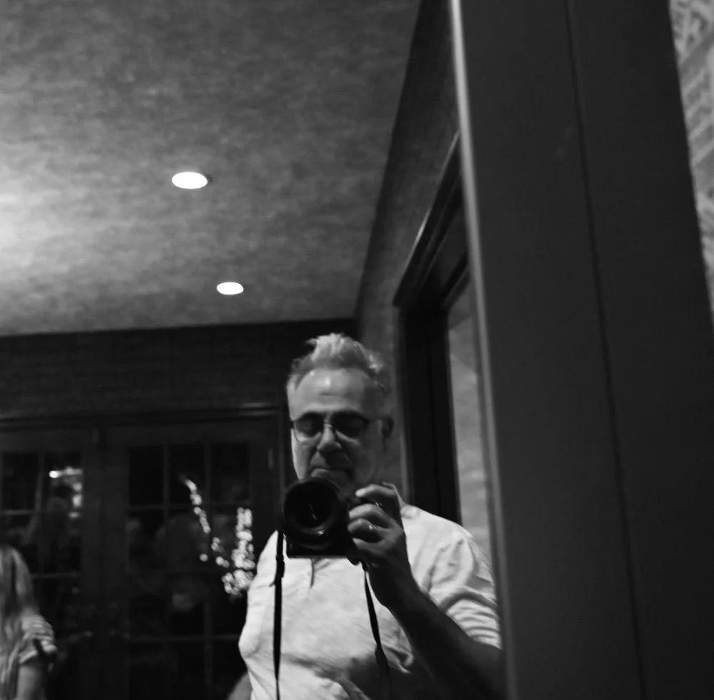 Black and white photo of a man taking a selfie in a mirror, wearing glasses and a light-colored shirt, in a room with wooden walls and ceiling.