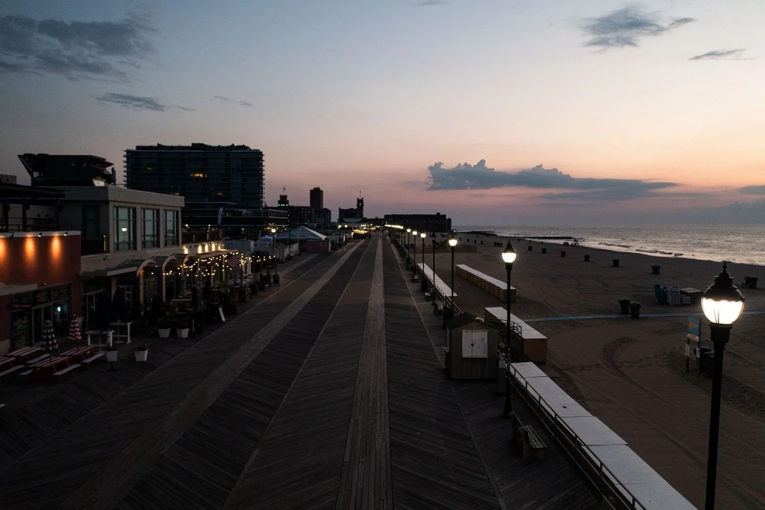 Empty boardwalk along the beach at dusk with street lamps lit and buildings in the distance.