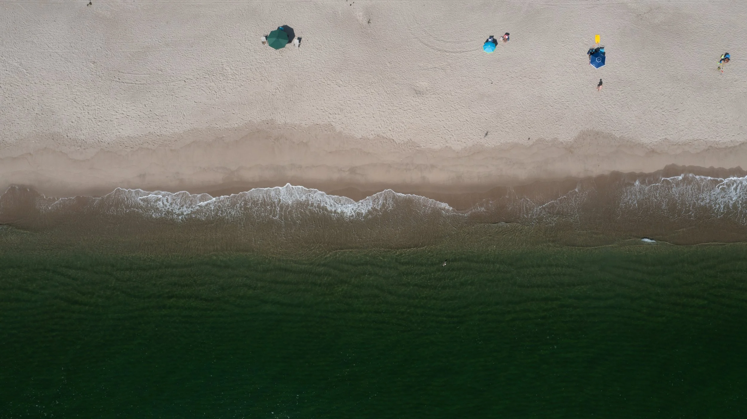 An aerial view of a beach with sand, ocean waves, and a grassy area. There are small umbrellas and people walking along the shore.