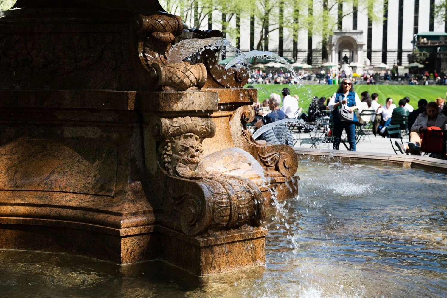Close-up of a historic ornate stone fountain with water flowing from spouts, with people sitting and walking in a park on a sunny day in the background.