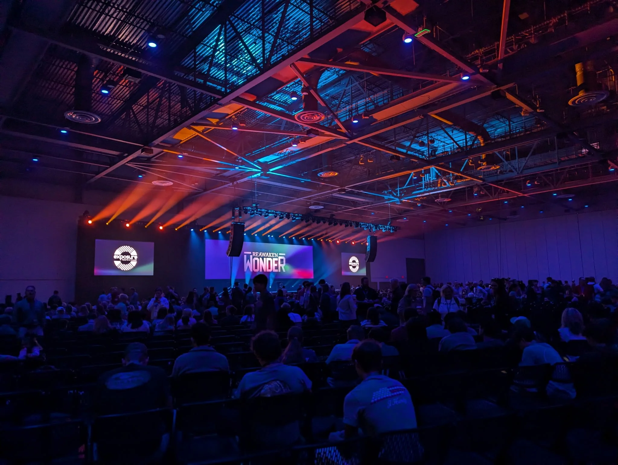 An indoor conference or event with people seated in front of a stage with lighting and screens displaying the event's name, 'Reawaken Wonder,' and the logo of Exposure Youth Camps.