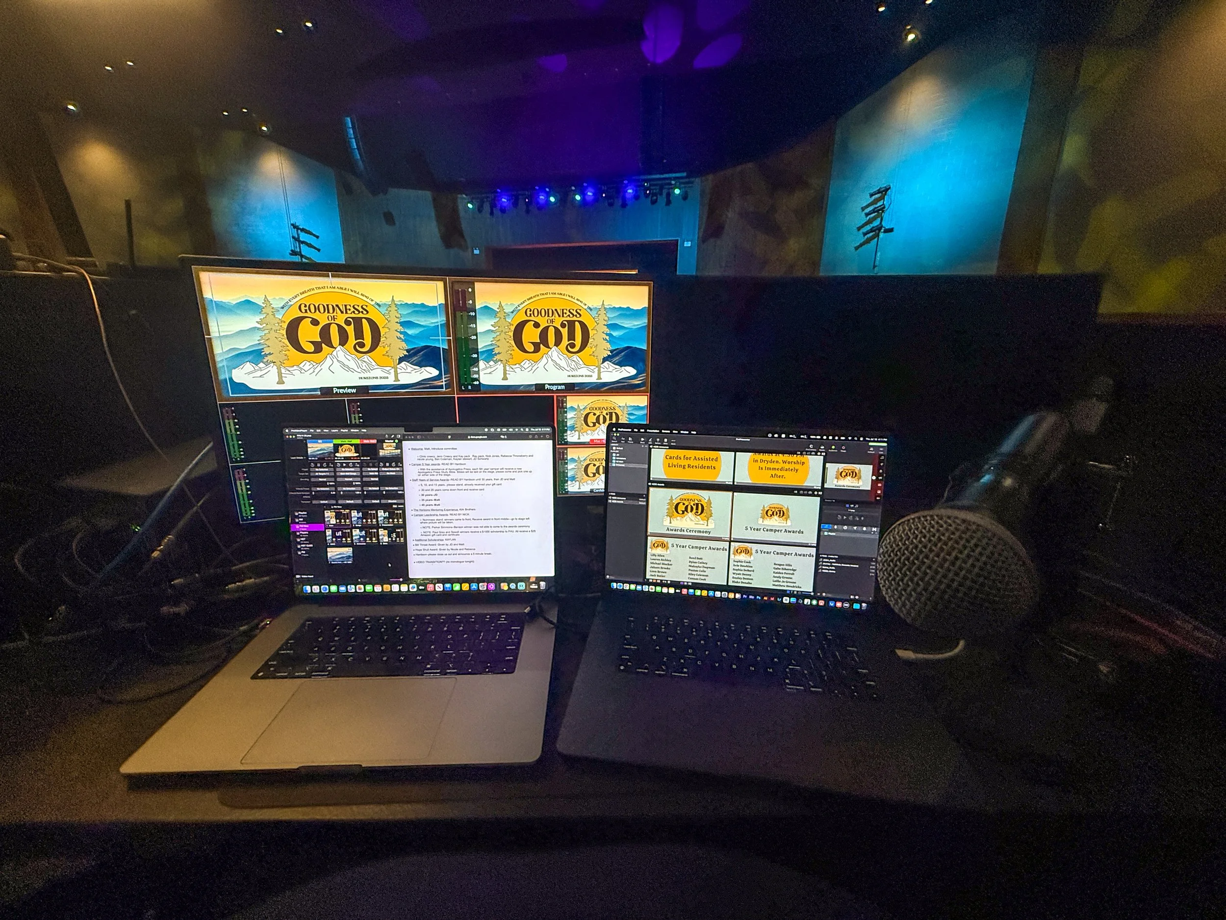 A control room or studio with multiple computer monitors displaying presentation slides for the event 'Goodness of God' with mountains and trees in the background.